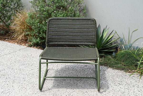 Green outdoor chair with woven design on a gravel surface with plants in the background
