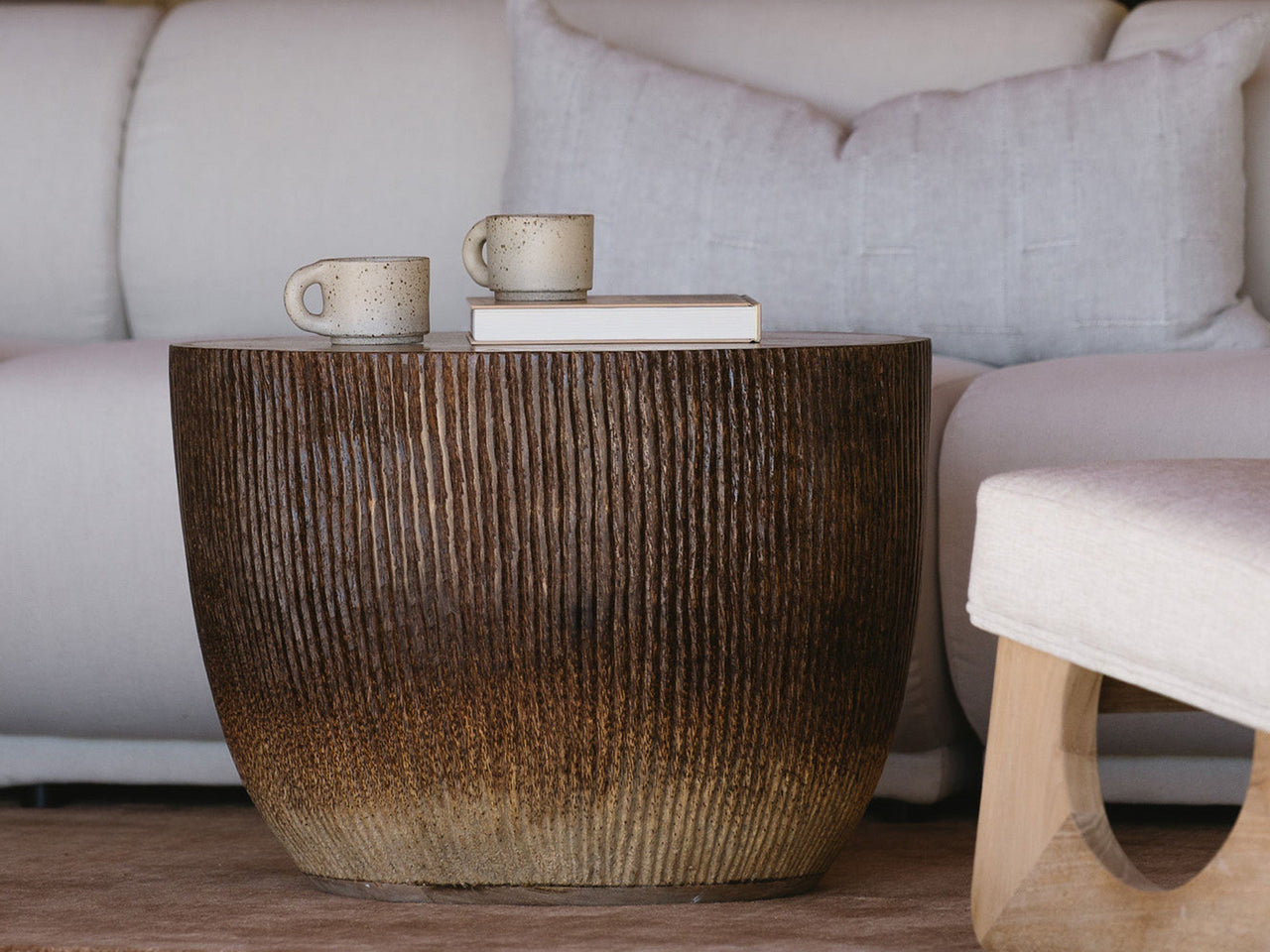 A round, ribbed dark brown wooden coffee table styled in a modern living room setting with a light beige sectional sofa in the background.