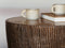 A close-up of a round wooden side table with a textured, bark-like vertical grain pattern. Two speckled ceramic mugs and a book rest on the tabletop against a soft, out-of-focus background.
