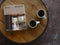 A book titled 'BUSH MODERN' by Thomas S. Bush lies on a round wooden slice tray, next to two white ceramic mugs filled with dark coffee, set against a dark textured background.