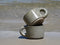 Two stacked ceramic mugs on a sandy surface with a blurred natural background